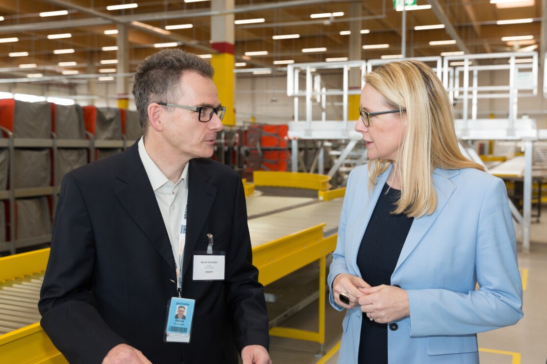 Ein Herr im Anzug und mit Brille unterhält sich mit einer blonden Dame, die ein hellblaues Sakko trägt. Im Hintergrund: ein Förderband.