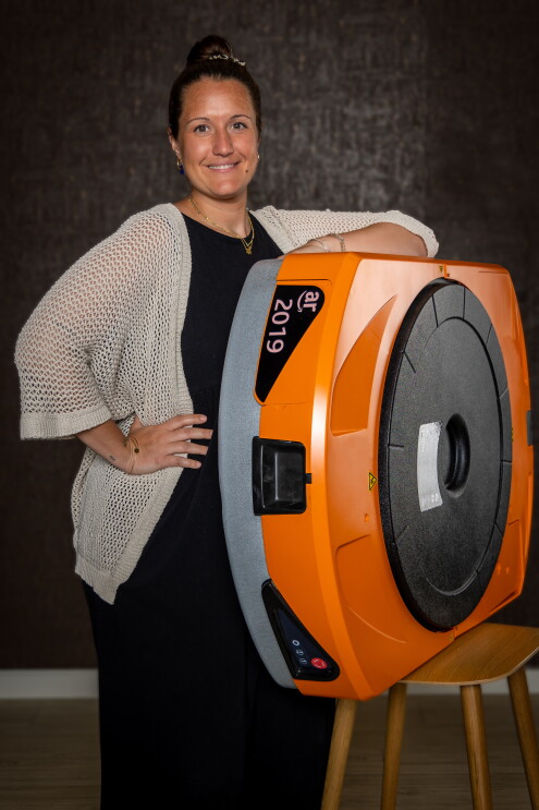Carmen Veuthey, responsable de Operaciones en el centro logístico de Illescas. El fondo es negro, encima de un taburete hay un robot de Amazon de color naranja. Carmen está apoyada con el brazo izquierdo al robot. Va vestida con un vestido negro largo y una chaqueta crochet de punto de media manga y hasta la cintura de color beige. La mano izquierda la tiene en la cintura. Va con collares de oro, pendientes y un moño alto. 
