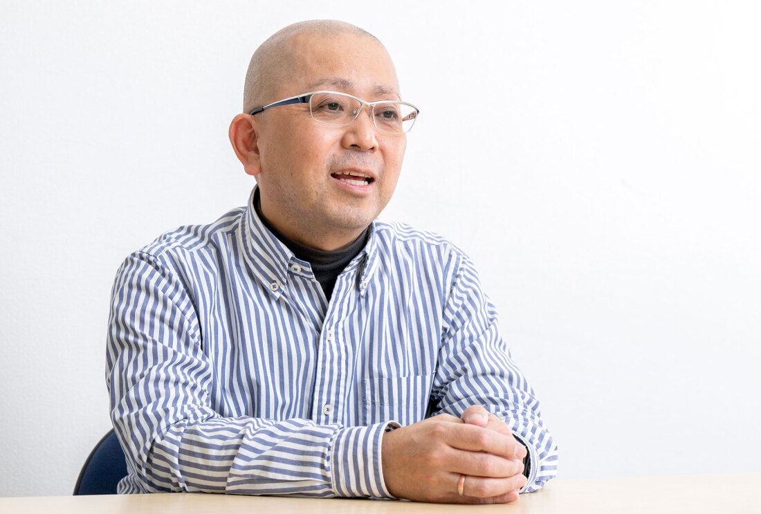 白い壁を背景に話をする人物。テーブルに向かって座り、両手を軽く組んでいる。薄い青と白のストライプシャツの下に黒いハイネックを着用し、眼鏡をかけている。