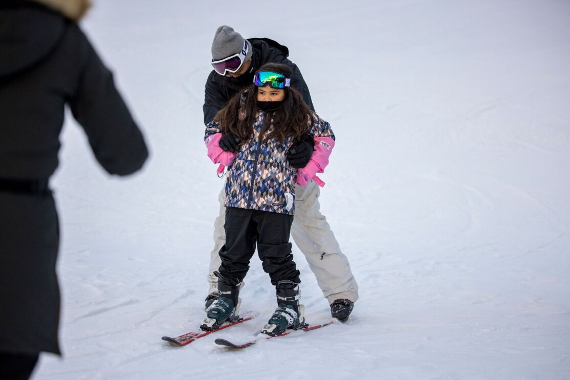 A father helps his young daughter down a ski slope on skis in the wintertime.