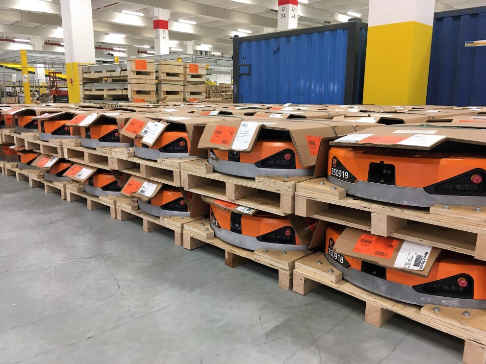 Auf Paletten stapeln sich orange-farbene Amazon Transportroboter. Sie ähneln haushaltsüblichen Saugroboter, sind aber deutlich größer.