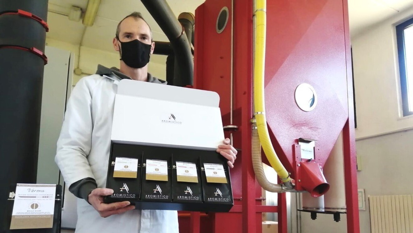 Un uomo in un sito di produzione di caffè indossa un camice bianco e una mascherina protettiva sul volto. Regge in mano un espositore di confezioni di caffè in polvere. 