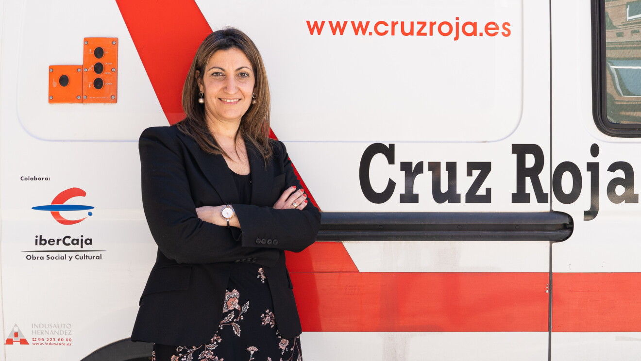 Olga Díaz, subdirectora de Inclusión Social de Cruz Roja. Con un vestido negro estampado con flores rosas, unas medias negras y una americana también negra. el pelo castaño liso por el hombro y uos pendientes con una perla colgando y un reloj en la muñeca izquierda. Está con los brazos cruzados apoyada en una furgoneta de Cruz  Roja. La furgoneta es blanca y lleva escrito Cruz Roja. 