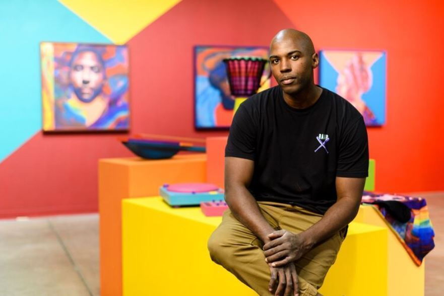 An image of Thomas Evans sitting on a yellow cube in front of a colorful art installation. 
