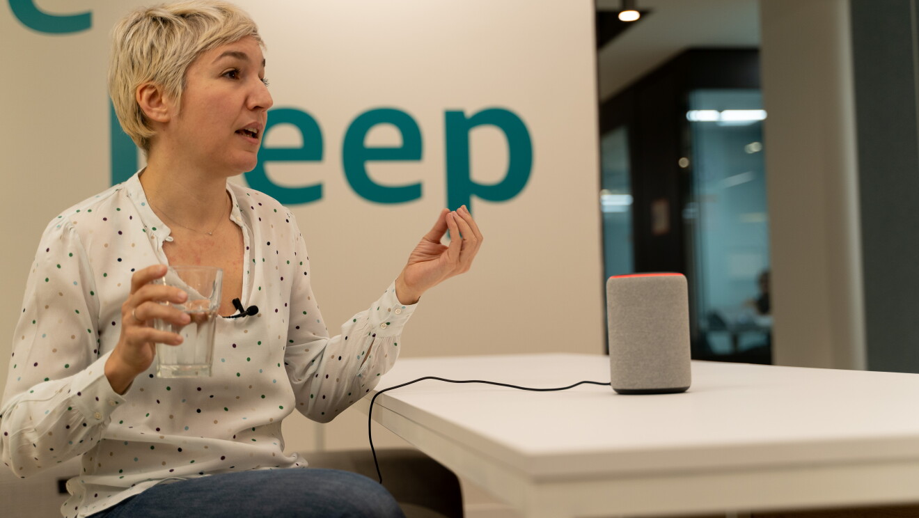 Victoria Pollo, Consumer Marketing Alexa Skills. Victoria, en la izquierda de la foto, con el pelo corto, rubo y una camisa blanca con lunares pequeños, está sentada y tiene un vaso de agua en la mano. Enfrente tiene una mesa blanca con un dispositvo Echo de color gris. 