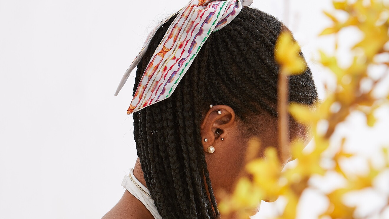 Primer plano de la cabeza de una persona racializada, con trenzas en el pelo y una cola atada con uno de los pañuelos de SOULEN, La mujer está mirando hacia abajo, tiene la oreja con cuatro piercings y en el cuello el tirante de un vestido blanca. Su cara queda detrás de unas hojas de color amarillo.