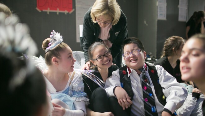 一个男孩和几个女生坐在一起在后台。他们穿着舞蹈服装。在后台,一个女人靠了下来。