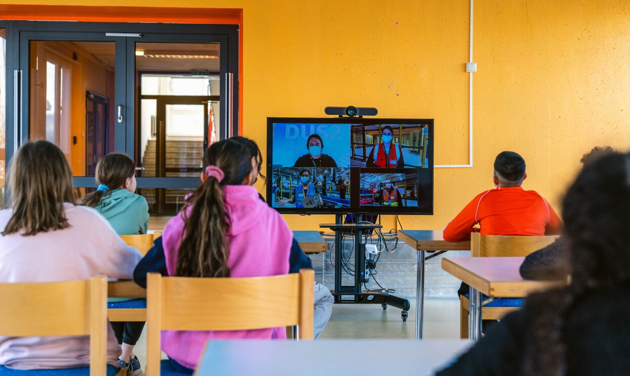 Kinder der Arche sitzen in einem Klassenzimmer und schauen auf einen Bildschirm. Dort stellen sich gerade die Tourleiter:innen der AFE-Tour vor.