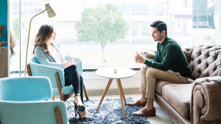 A man sits on a couch across from a woman therapist who is sitting in a blue chair and writing on a notepad.