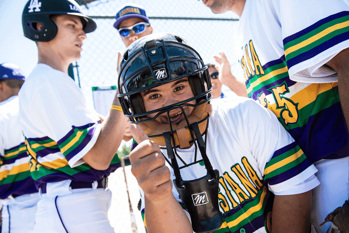 An image of a person wearing a baseball helmet and uniform giving a thumbs up. The background shows other people in the same uniform doing a handshake.