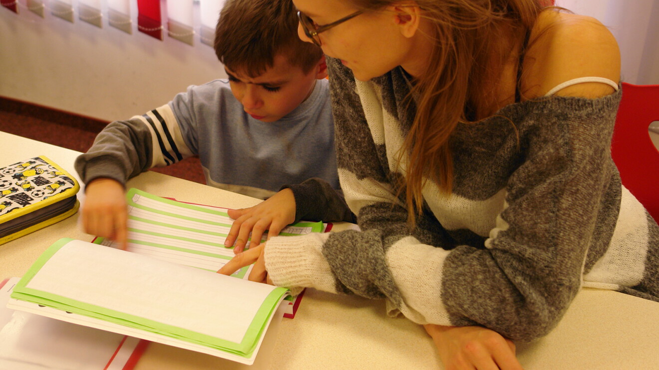 Ein kleiner Junge und eine junge Frau sitzen gemeinsam an einem Tisch und schauen auf ein Schulheft.