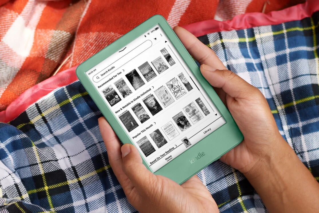 赤と青のタータンチェック柄の毛布の上で小さな緑のタブレット(Kindle)を両手で持っている人の手。スクリーンには本の表紙が並ぶKindle ストアが表示されている