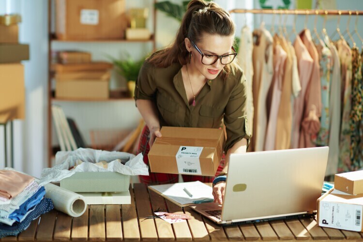 Eine Frau mit Brille steht vor einem Laptop und hat ein Paket in der Hand.