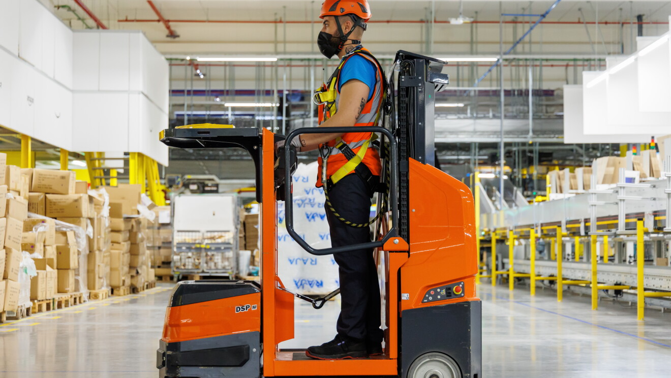 En la entrada de un centro logístico de Amazon aparece un asociado subido a un toro mecanizado. Rodeado de palés con productos y una cinta transportadora. El empleado va con casco y botas de seguridad, chaleco, camiseta de manga corta y pantalón largo y un arnés atado al toro. Las manos al volantes y mirando al frente. 