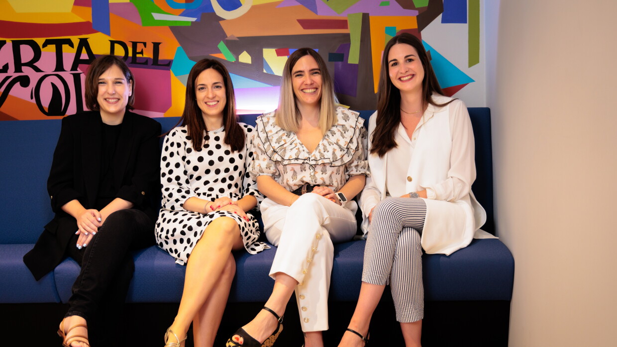 Sentadas en un sofá de color azul y con un fondo pintado de muchos colores vivos y las letras en negro de Puerta del Sol, están sentadas cuatro mujeres con la pierna izquierda cruzada. De izquierda a derecha: María Alonso vestida con un traje pantalón de color negro, con un jersey negro también debajo de la americana. Lleva unas sandalias marrones con dos tirantes. Al lado está Alejandra Ossa tiene el pelo negro largo hasta el hombro. Lleva un vestido blanco de lunares negros de manga larga y largo hasta la rodilla.  Lleva unas sandalias de color de oro de plata. Lara Fajardo es la cuarta de la foto. Va con una camisa blanca estampada, con el pelo hasta el hombro con mechas negras y decoloradas hasta el rubio en las puntas. Con pantalones blancos y unas sandalias negras de plataforma de color de mimbre de color marrón y negro. Y, finalmente, Miriam Domínguez-Adame Aguilera con el pelo liso negro hasta el pecho. Unas sandalias negras, unos pantalones rayados de color negro y blanco, una camisa y chaleco de color blanco.
