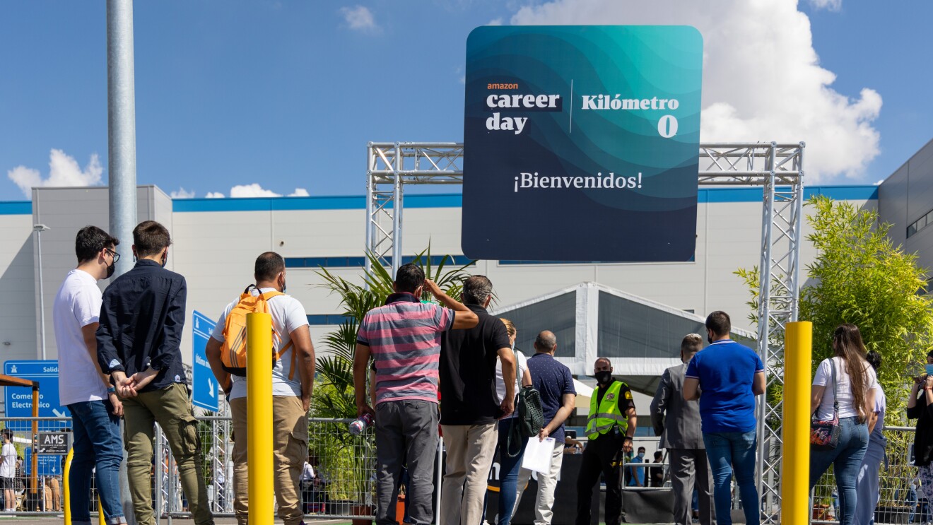 30 000 personas solicitaron un puesto de trabajo en Amazon durante el Career Day. En primer plano un grupo de 15 personas de espaldas a la cámara haciendo cola para participar en el Career Day de Amazon 2021. De fondo el centro logístico de Amazon en Illescas. La gente está haciendo cola, en la entrada, delante de un cartel que está apoyado en la parte superior de una columna, En el cartel pone ¡Bienvenido!, kilómetro 0 y Amazon Career Day. Se ve el cielo azul con nubes blancas.