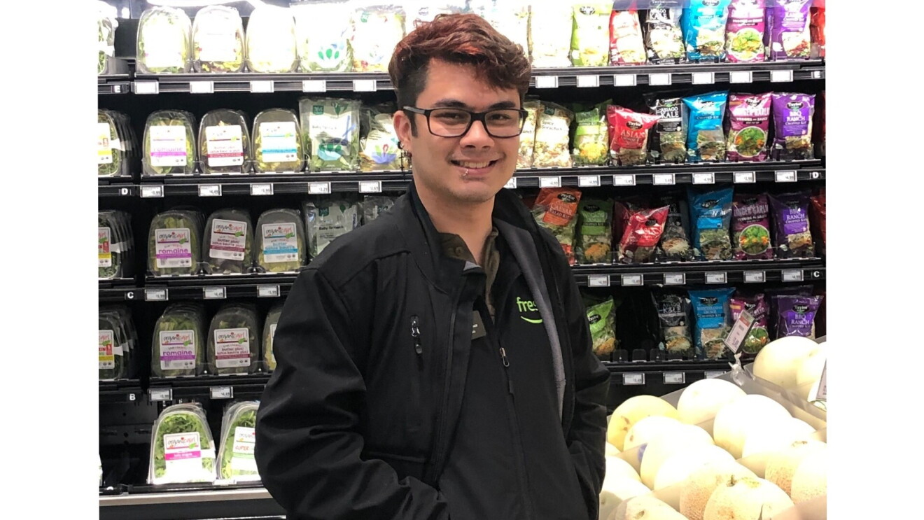 Eli smiles for the camera in front of the packaged salads and produce section of the grocery store. He wears a black Amazon Fresh zip-up and black-framed eyeglasses.
