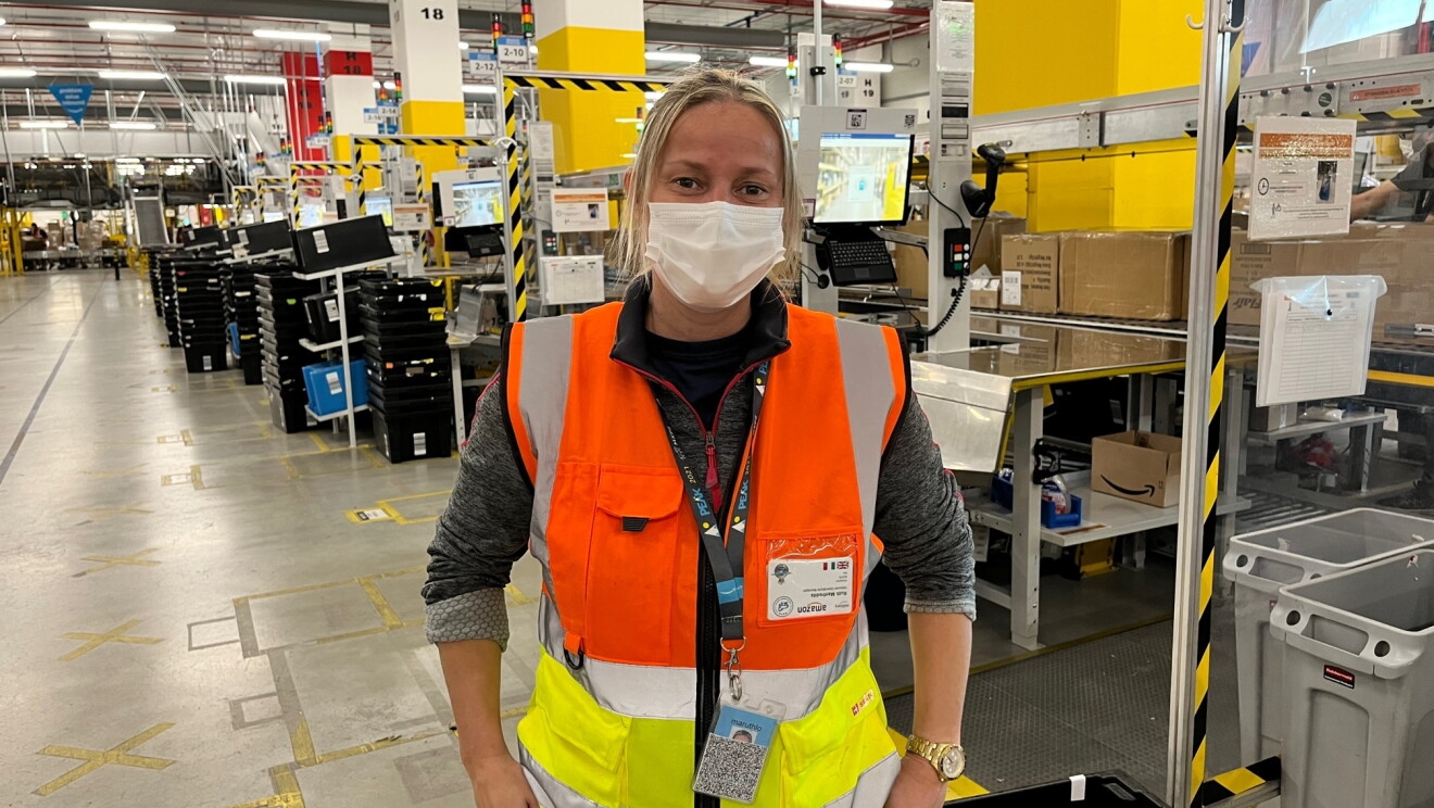 Mezzo busto di una dipendente Amazon all'interno di uno dei magazzini. Ha le maniche arrotolate, i capelli legati e indossa una mascherina chirurgica sul volto 