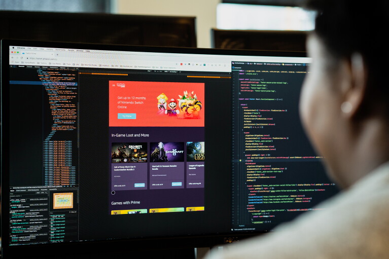 A woman sits at a computer, with two monitors in front of her. She is working within software, as illustrated by the lines of code displayed on the monitors.