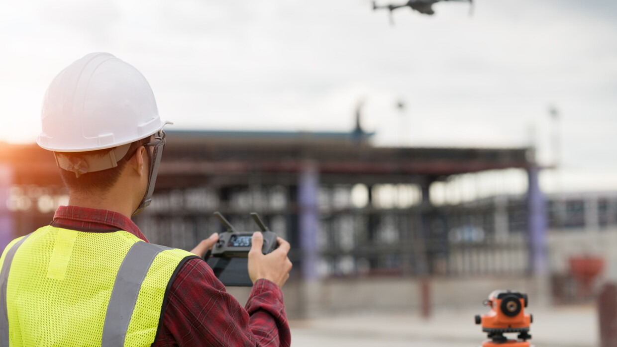 A man wearing a hardhat and yellow safety vest maneuvers a drone at an Amazon fulfilment centre site. 