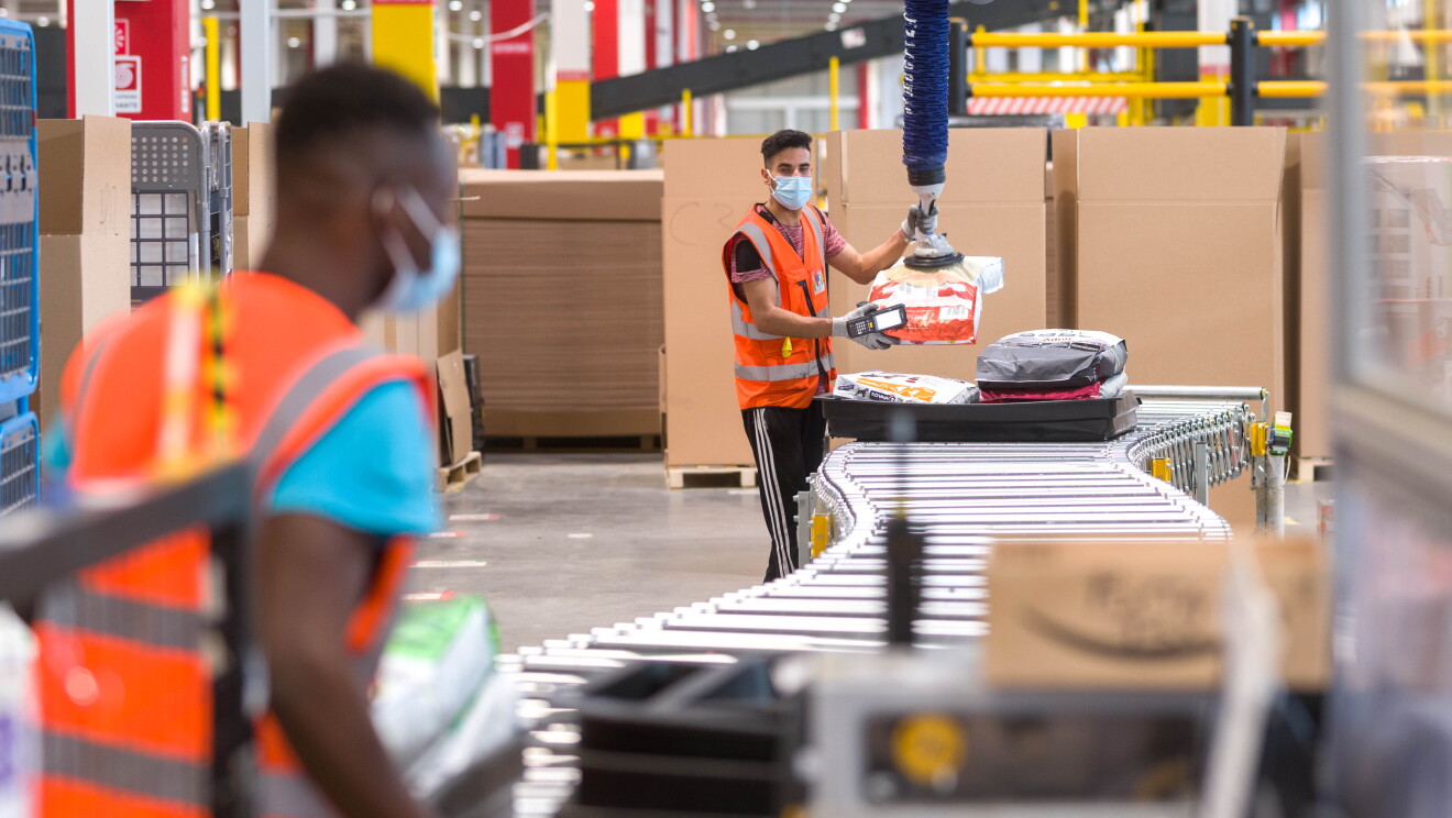 Empleados de Amazon que se benefician de una herramienta que ayuda a dejar objetos más pesados (también conocida como elevador por vacío). Dentro de un centro logístico hay dos asociados delante de una cinta transportadora. De fondo se ven cajas muy grandes de color marrón y cintas transportadoras. El asociado que es´tá en primer plano tiene el pelo muy corto, lleva una mascarilla quirúrgica, un chaleco naranja y una camiseta azul. Está desenfocado pero se aprecia que está cargando, con las dos manos, paquetes. El otro asociado está al final de la cinta transportadora y agarra con una mano un paquete y la otra en el elevador por vacío. Va con pantalón negro, camiseta negra manga corta, chaleco naranja y guantes en las manos y en la derecha un lector de código de barras. 