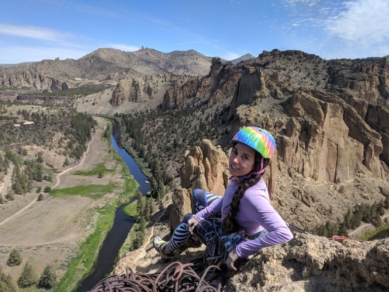 Marta is wearing rock climbing gear as she sits at the top of a high peak.