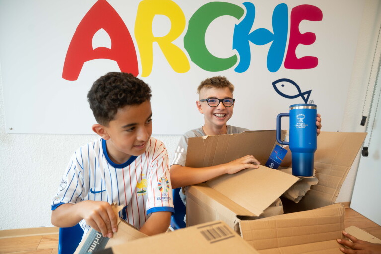 Zwei Kinder packen Pakete aus und stehen vor der Aufschrift "Arche".