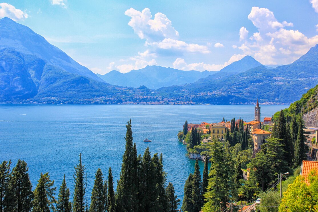 An image of a blue lake surrounded by green trees and tall mountains. There are yellow and oranges villas lining the shore of the lake.