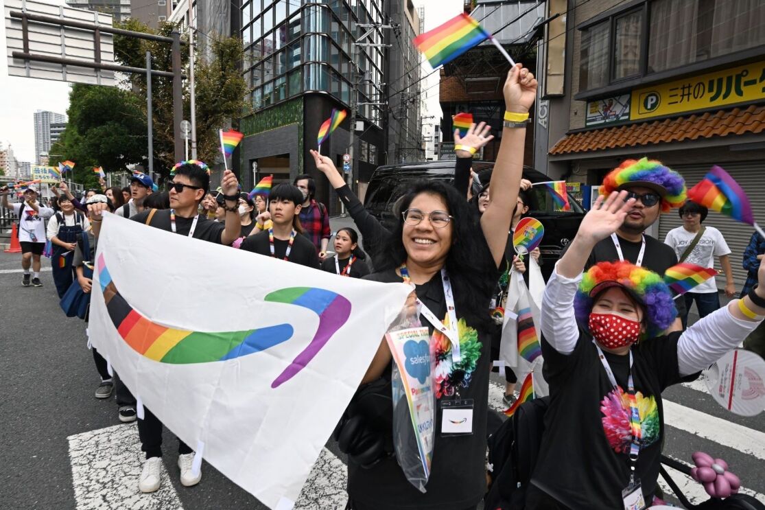 道で数人の人々が小さなレインボーフラッグを振っている。メガネをかけている女性とサングラスをかけている男性が虹色のAmazonのスマイルロゴの横断幕を持っている