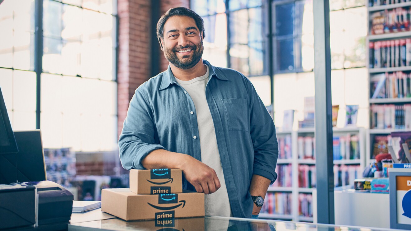 A business owner stands behind a desk and smiles with his arm resting on two Amazon packages.