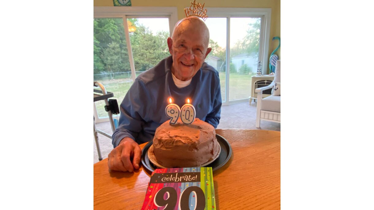 An image of Kate's dad at his birthday party. He is wearing a headband that says "Aged to perfection" and there is a cake in front of him with candles in the shape of the number "90." There are also colorful napkins in front of the cake that say "celebrate 90."