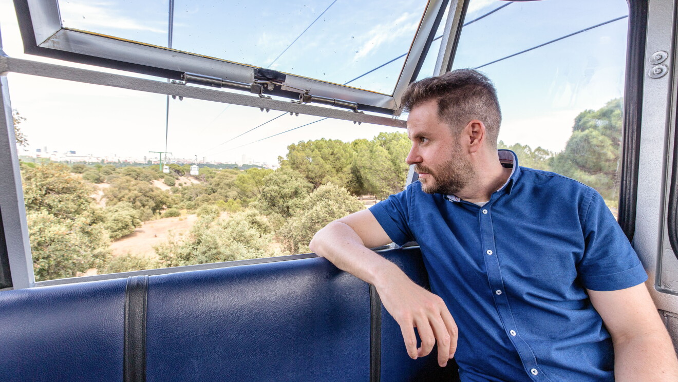 En el teleférico de Madrid, Paul Pen mirando al horizonte. La ventana de la cabina está abierta y  Paul tiene un brazo reposando en la silla. El escritor lleva una camisa azul y el pelo corto. 