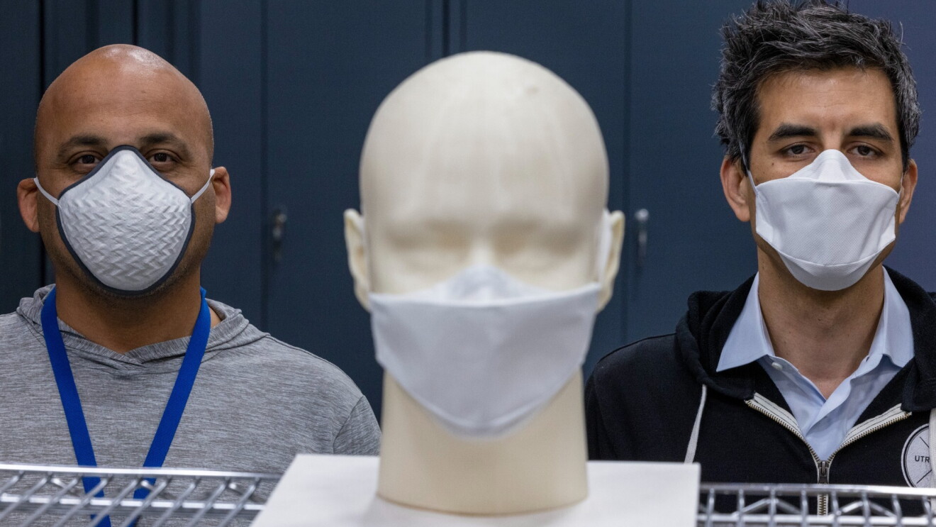 Two men wearing face masks stand behind a mask prototype.