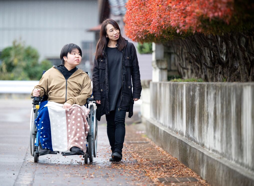 車椅子に座っている男性と黒いジャケットを着ている女性が道で散歩している。道のそばに赤くなっている木の葉が植えられて並んでいる。