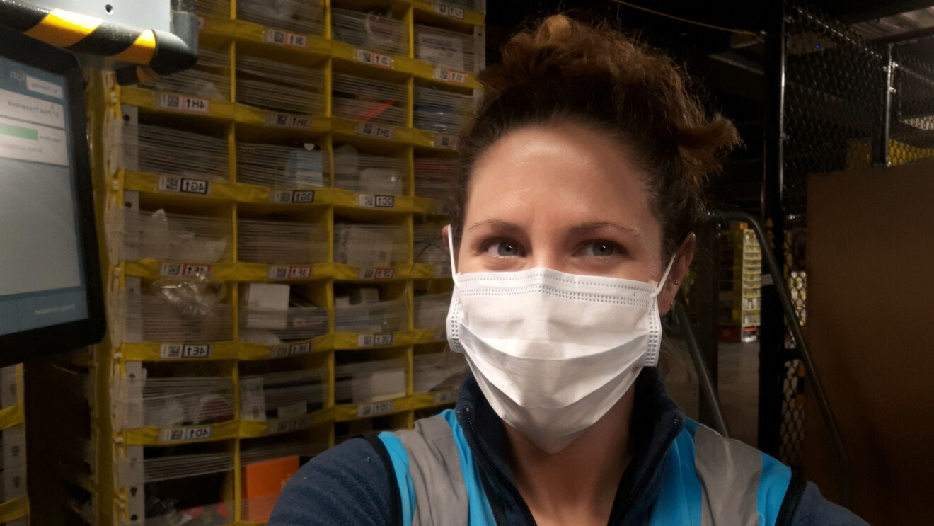 A woman in a safety mask stands with shelving behind her.