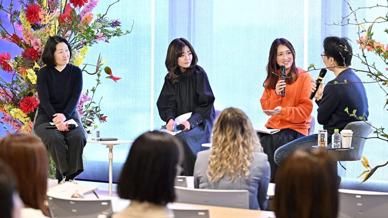 3人の女性と男性のMCはステージで話している。彼女たちの後ろには大きな花が置かれている。