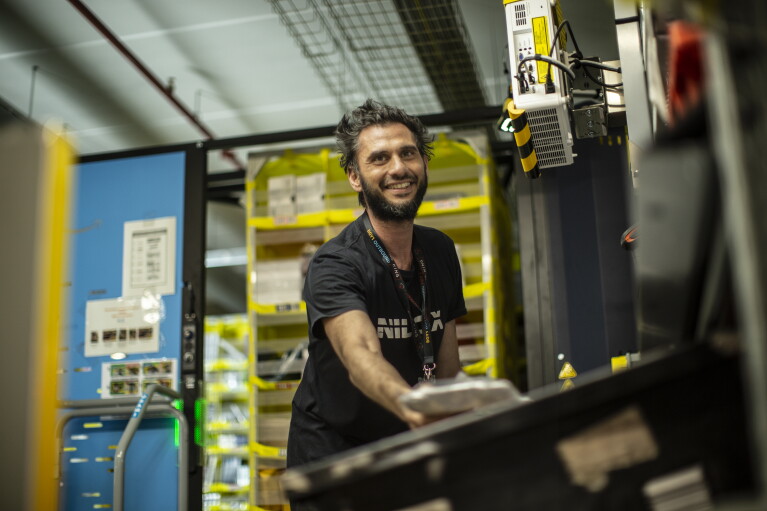Elia works in a FC. He has a black tshirt, a big smile and he is picking an item. ELIA VILLARDITA - Operatore di magazzino :"Sicurezza sul lavoro significa creare un ambiente lavorativo dove non ci sono condizioni di pericolo. Avere sicurezza sul lavoro porta tranquillità, è bello sapere che i rischi sono quasi nulli, questo ci permette di lavorare meglio e quindi in modo più produttivo".
