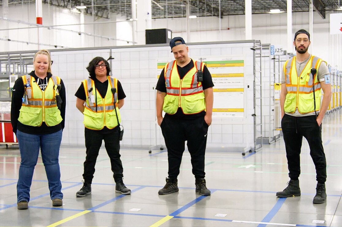 Four Amazon Operations employees wear yellow safety vests and stand and smile in a fulfillment center.