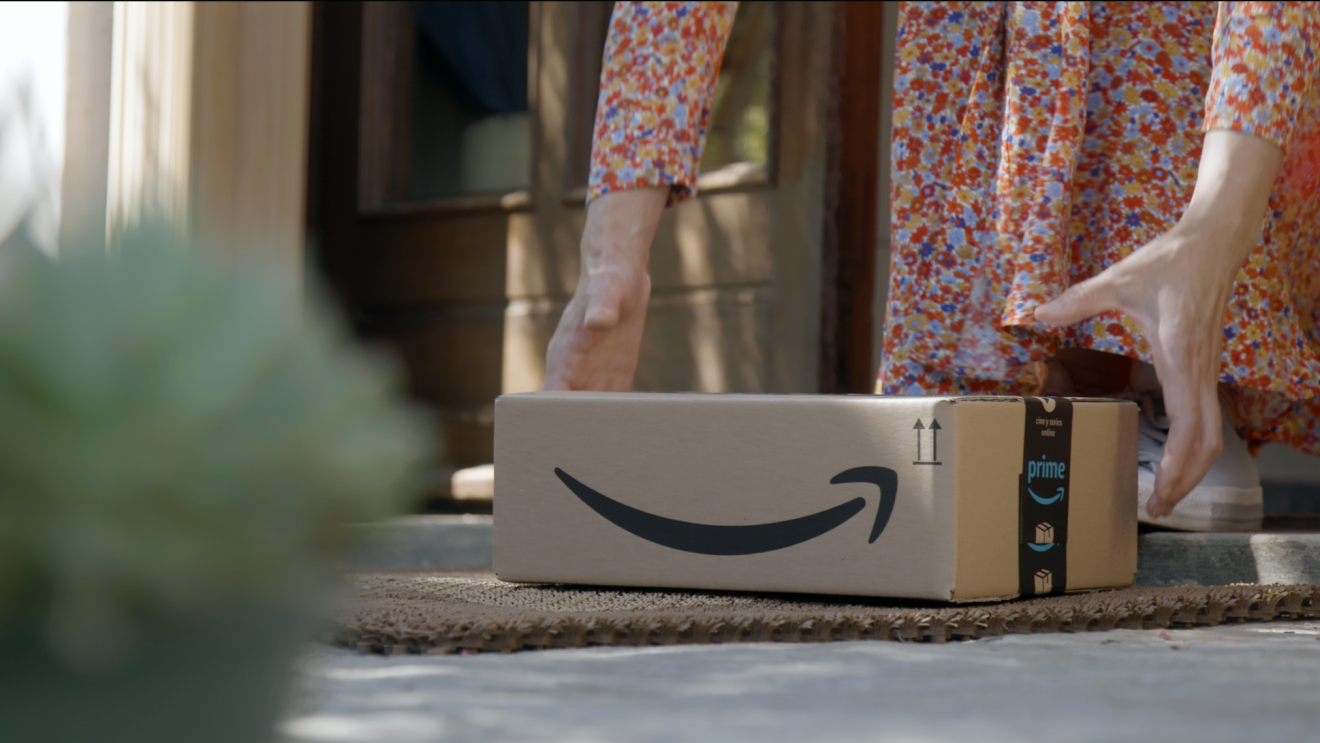 Scatola Amazon in primo piano, posata su uno zerbino di fronte all'ingresso di una porta. Si vedono due mani di una donna che stanno per raccogliere il pacco. La donna indossa una gonna a fiori e delle sneakers. 