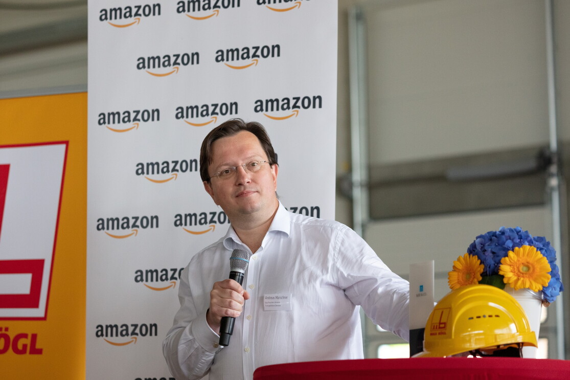 Andreas Marschner, Vice President Transportation Services Europe bei Amazon Andreas Marschner mit einem Mikrophon vor einem Stehtisch. Im Hintergrund eine Fotowand mit Amazon Logos.