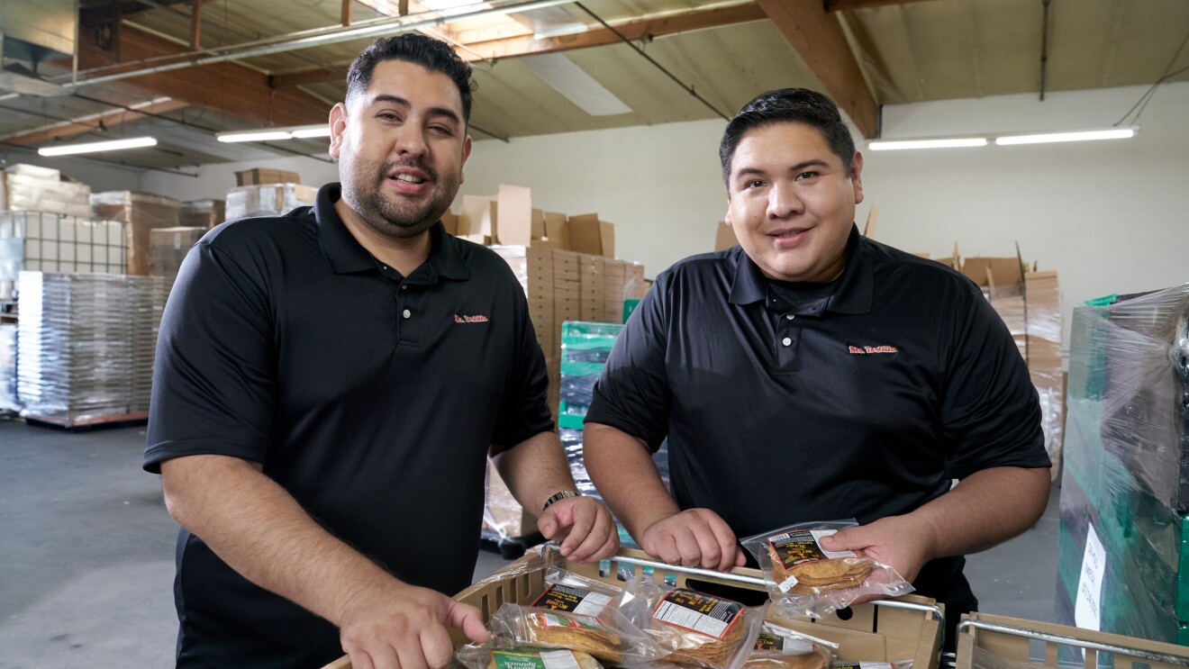 Anthony and Ronald stand side-by-side leaning on pallets of their packaged tortillas.