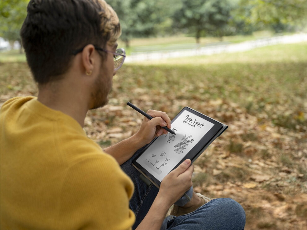 Ein Mann mit Brille und gelben Pullover sitzt in einem Park. Er hält den neuen Kindle Scribe in der Hand, mit welchem er Bilder in sein "Garten Tagebuch" schreibt.