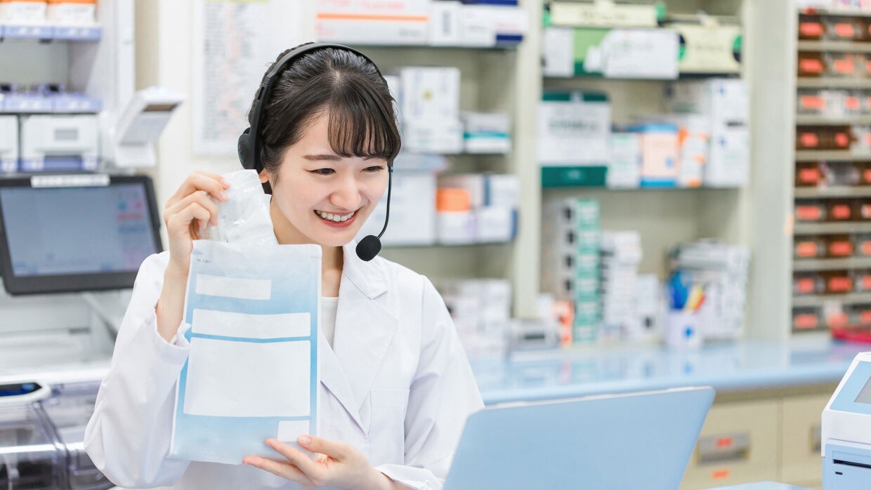 薬棚の前で、ヘッドセットを着けた白衣の女性薬剤師がカウンターに置いたノートパソコン画面に向かって処方薬を袋から取り出し見せている