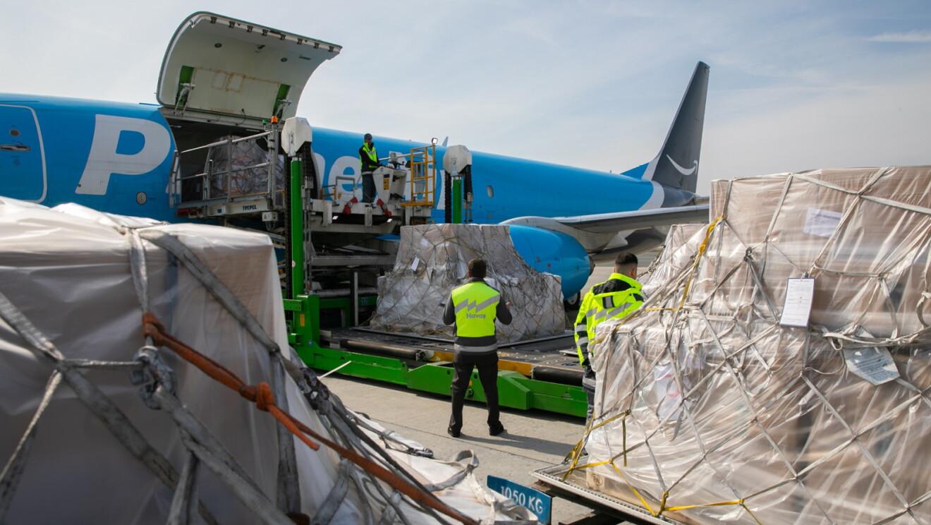 En la pista de un aeropuerto hay cuatro palés cargados con ayuda humanitaria. Los palés son de papel marrón, con pegatinas blancas y unas cuerdas. De fondo un avión de color azul con las letras en blanco Prime Aire. El cielo está nublado y es el atardecer. Los palés están arrastrados por una camioneta verdey el avión tiene uan grúa para cargar la ayuda. En la foto aparecen tres personas con chaleco amarillo. Una persona encima de la grúa y las otras dos en la pista. 