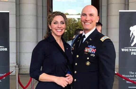 An image of a couple smiling for a photo outside of a building with red ropes in front of it. The man is wearing a military outfit and there are signs for Wounded Warrior Project behind them.