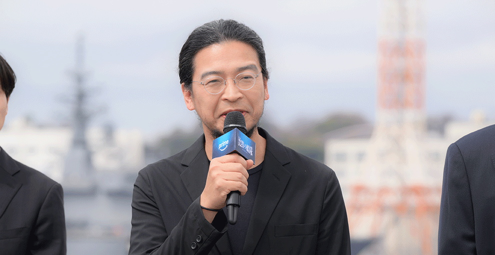 日中の屋外で、黒のジャケットを着てマイクを持った男性。口にマイクを当て話している。背景に白と赤の電波塔のようなものが見える。