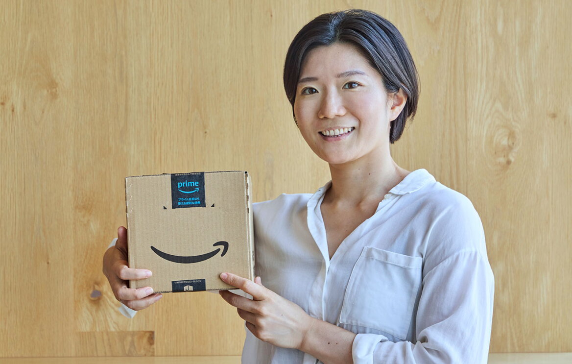 室内で、木目の壁を背にして笑顔を見せる女性。小脇にAmazonロゴ入りの小箱を抱えている。