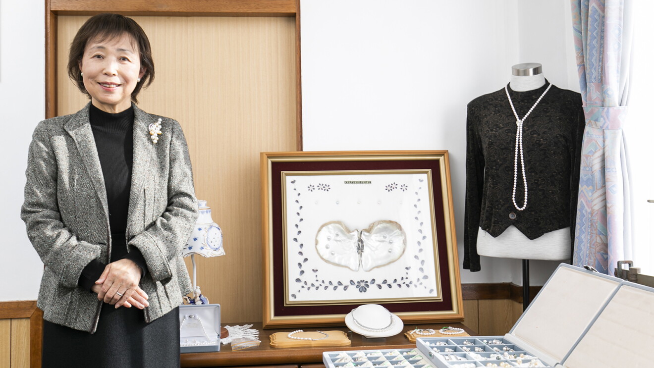 白い壁の室内、グレーのジャケットと黒いワンピースの女性が立っている。女性の右側にさまざまな真珠製品のディスプレー。