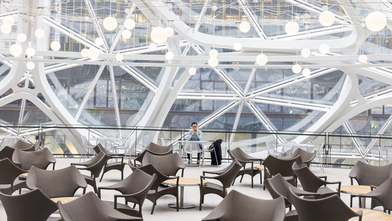 An image of several sets of chairs and tables. There is a man sitting toward the back of the photo looking at his phone. The wall behind him is all made of glass windows.  