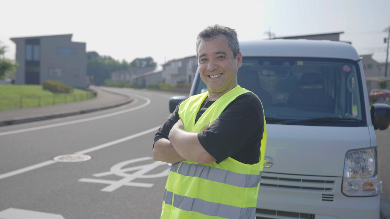 黄色の安全ベストと黒いTシャツを着ている男性（国吉 ルイスさん）。住宅地の近くに止められた軽自動車の前で腕を組んだポーズとしている